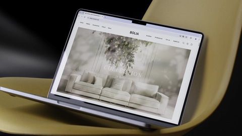 Laptop viser Bolia's hjemmeside med fokus på 'Spring Collection' og et stilrent sofa-design, placeret på en gul stol.